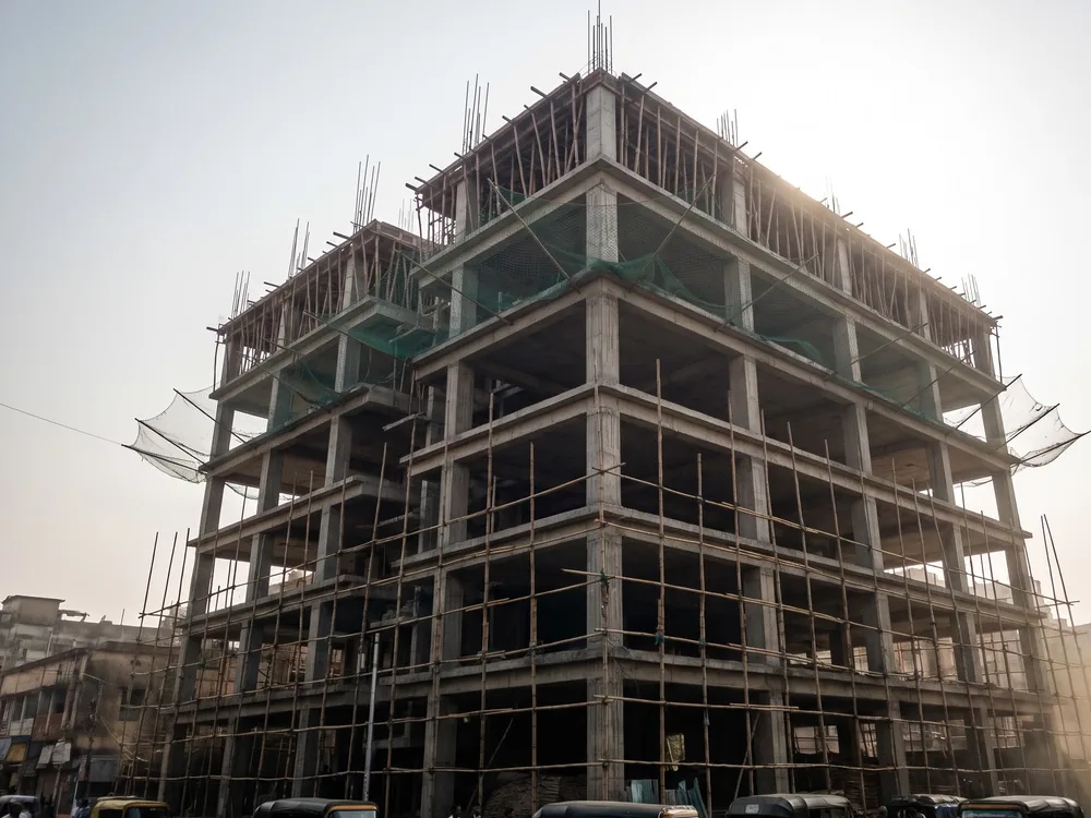 A concrete-framed building under construction, wrapped in bamboo scaffolding and green safety netting, autorickshaws parked at street level.