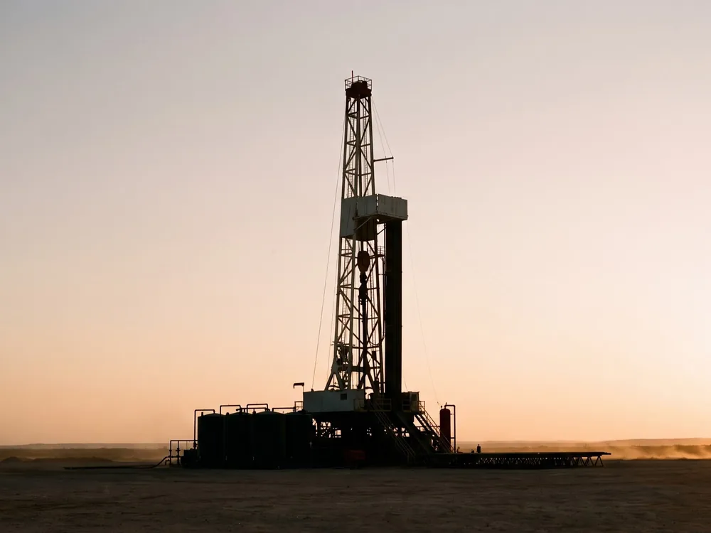 An onshore drilling rig silhouetted against a pale dawn sky on a flat dusty plain.