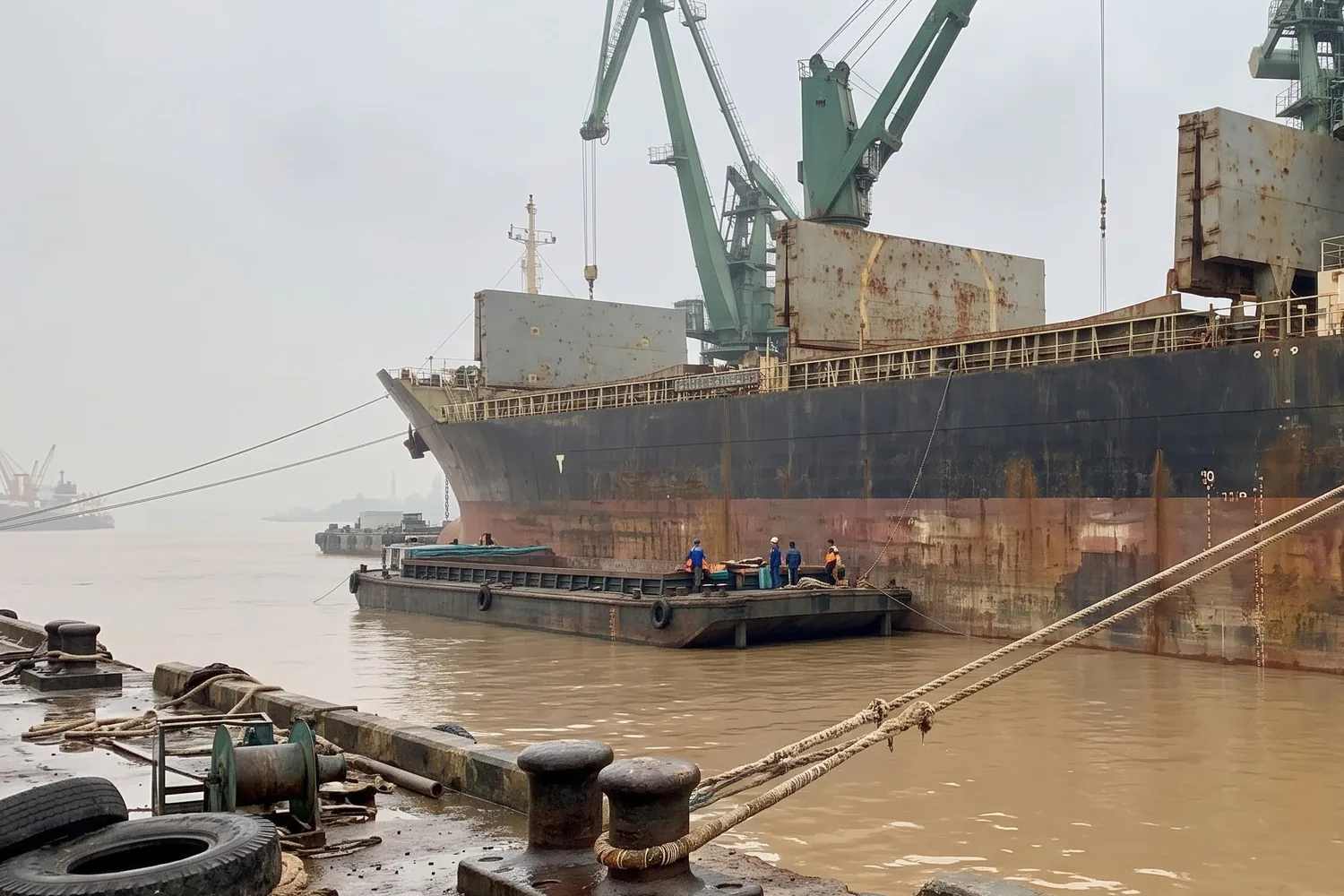 A rust-streaked cargo ship at berth under green quayside cranes on a muddy brown river, mooring lines running to bollards in the foreground.