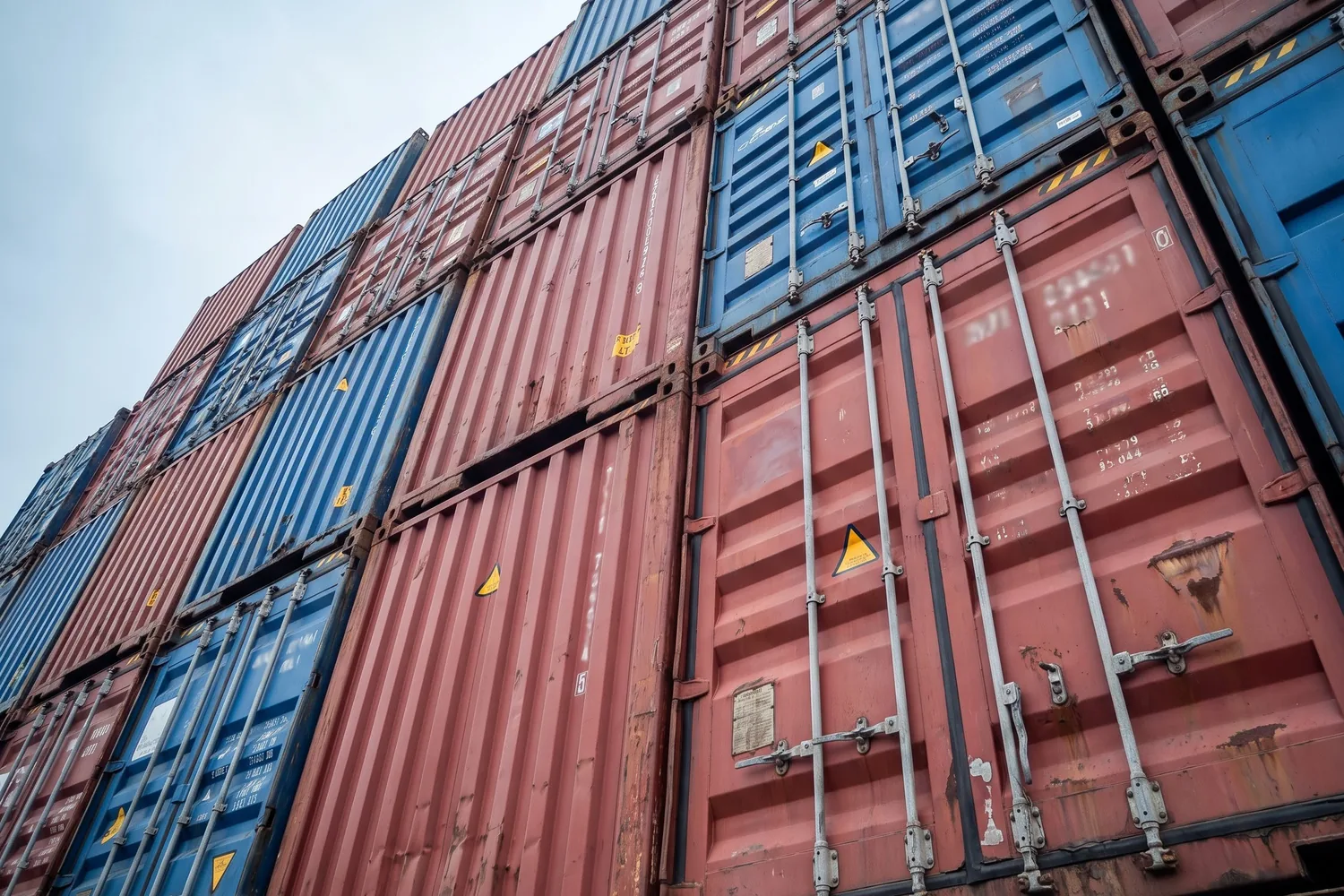 Stacked shipping containers in weathered oxide red and blue, shot from below so the rows run diagonally across the frame.