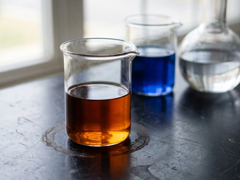 Plain glass beakers on a scuffed black laboratory bench holding amber, cobalt blue and clear liquids.