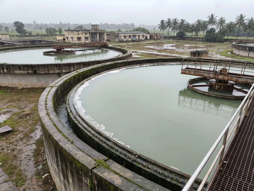 Circular concrete clarifier tanks with rotating scraper bridges at a water treatment works, under monsoon overcast, paddy fields and palms beyond.